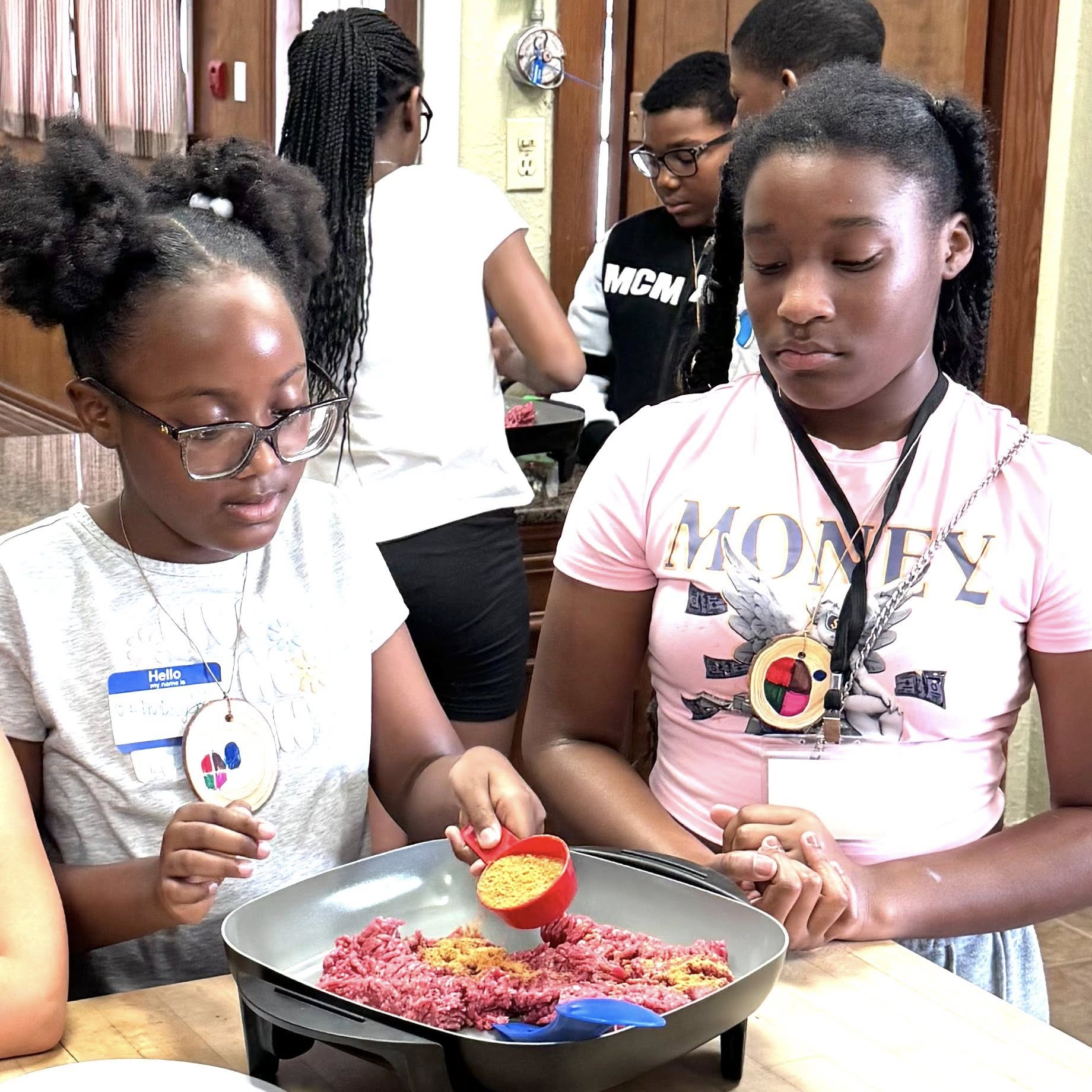 One girl watching another girl pour seasoning over meat in a skillet.